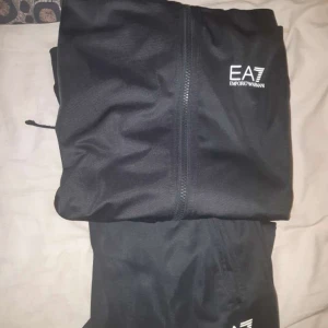 Svart sett från Emporio Armani EA7 originalen  - Svart jacka från Emporio Armani EA7 med dragkedja framtill och logga på bröstet. Snygg och stilren design, perfekt för dig som gillar klassiska streetwear-vibbar. Jackan har en enkel look och är lätt att matcha med olika outfits.