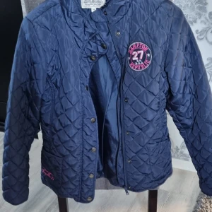 Marinblå quiltad jacka Hampton Republic - Quiltad marinblå jacka från Hampton Republic med snygga detaljer som tryckknappar, dragkedja och broderad logga på bröstet. Jackan har två fickor fram och är perfekt för kyliga dagar. Klassisk och sportig look med lång ärm.