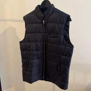 Marinblå quiltad väst från John Henric - Snygg marinblå väst med quiltad design och hög krage. Västen har två fickor framtill med dragkedja och en liten bröstficka. Perfekt att slänga över en hoodie för extra värme. Stängs med hel dragkedja framtill. Märke: John Henric 