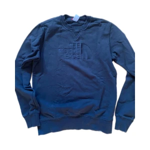 Mörkblå sweatshirt från The North Face - Snygg svart sweatshirt från The North Face med broderad logga framtill och bak på axeln. Tröjan har rund hals, ribbade muddar och är tillverkad i mjuk bomullsmix. Perfekt för en chill och stilren look.