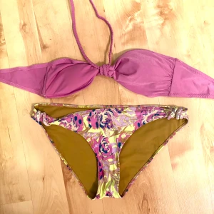Lila och blommig bikini med knyt - Snygg bikini med lila bandeau-topp som knyts i nacken och har spänne bak. Underdelen har blommigt mönster i lila, gult och inslag av blått. Materialet är mjukt och stretchigt, perfekt för stranden eller poolen.