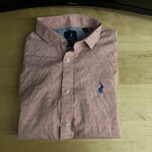 Rosa skjorta från Polo Ralph Lauren - Rosa skjorta från Polo Ralph Lauren med små diskreta mönster och klassisk blå broderad logga på bröstet. Skjortan har lång ärm, knappar framtill och är tillverkad i mjuk bomull. Perfekt för dig som gillar stilrena detaljer och snygg passform.