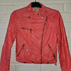  skinnjacka från Softy, stl S - Snygg och färgstark bikerjacka i korall/rosa från märket Softy Outerwear collection. Jackan har flera dekorativa dragkedjor och knappar som ger en cool stil. Färgen gör sig rättvisast på bilderna.  OBS! Jackan har vissa skavanker: *Slitage i nacken *Mindre repa på ena ärmen  *Märket inuti är något slitet *Reva på axlarna efter att ha hängt på galge i garderoben i 2 år * mörkare märken på armarna och lite på axeln. 