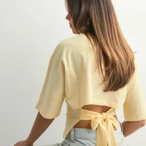 Gul omlottopp med knyt i ryggen - Snygg ljusgul topp med trekvartsärm och öppen rygg som knyts med en stor rosett. Knappt använd och inga defekter 