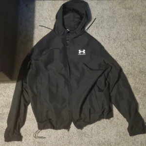 Svart vindjacka från Under Armour helt ny  - Svart vindjacka från Under Armour med huva och dragsko nertill. Jackan har dragkedja framtill och logga på bröstet. Tillverkad i lätt polyester, perfekt för blåsiga dagar. Snygg och sportig look med lång ärm och enkel design. Den är helt ny köpte den för en vecka sen men den var för stor för mig och råkade dra väck prislappen så kunde inte skicka tillbaka den det är därför jag säljer den..  buda på den kan tänka mig gå ner i pris... och bilderna är lite suddiga det är min kamera som är sönder.. 