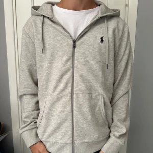 Grå hoodie från Polo Ralph Lauren - Snygg ljusgrå hoodie från Polo Ralph Lauren med dragkedja framtill, huva med snörning och klassisk broderad logga på bröstet. Tröjan har två fickor och är tillverkad i mjuk bomullsmix. Perfekt för en chill och stilren look.