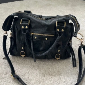 Svart city väska med gulddetaljer - Snygg svart axelväska i skinnimitation med guldfärgade detaljer och dragkedjor. Väskan har flera fack, både utvändigt och invändigt, samt justerbar axelrem och handtag. 