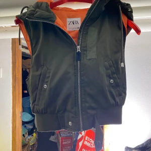 Grön dunväst från Zara med huva - Snygg mörkgrön dunväst från Zara med orange foder och huva. Västen har dragkedja framtill, två fickor med tryckknappar och ribbade muddar nedtill. Perfekt för lager-på-lager och streetstyle.
