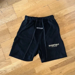 Svarta shorts Essentials Fear of God - Svarta shorts från Essentials Fear of God med broderad logga framtill. De har elastisk midja med snörning och en bakficka. Sköna och stilrena, perfekta för chill eller streetwear. Materialet är mjuk bomull och passformen är loose.