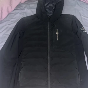 Svart Sail Racing dunjacka med huva - Svart dunjacka från Sail Racing med huva och dragkedja framtill. Jackan har Sail Racing-logga på bröstet och ett Sail Antarctica Expedition-märke på ärmen. Snygg, stilren och perfekt för kalla dagar. Fodrad insida och praktiska fickor med dragkedja.