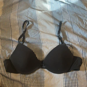 Svart vadderad bh med rosett - Svart bh med vadderade kupor och justerbara axelband. Söt liten rosett framtill mellan kuporna och klassisk hysk- och hakknäppning bak. Enkel och stilren design som passar till det mesta. Pris kan diskuteras.