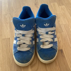 Blå adidas Campus sneakers i mocka - Snygga adidas Campus sneakers i blå mocka med vita detaljer och klassiska tre ränder på sidorna. Skorna har vit sula, rund tå och vita skosnören. Perfekta för dig som gillar retrostil och streetwear Skosnörena är knutna på ett annat sätt på bilden men har ändrat på de nu i efterhand!.