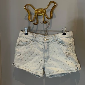 Ljusblå jeansshorts med nitar/bling - Gina Tricot - Snygga ljusblå jeansshorts från Gina Tricot med slitna detaljer. Shortsen är dekorerade med små silvriga nitar över hela framsidan. Klassisk femficksmodell och låg midja, perfekta för sommaren! 🍒☀️