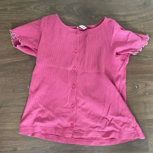 Rosa tröja  med glitter  - Supersöt rosa ribbad blus från Cubus med korta volangärmar och knappar framtill. Ärmsluten har en vågig kant med glittrig silvertråd som detalj. Perfekt för dig som gillar färg och vill ha en chill men ändå söt look. 