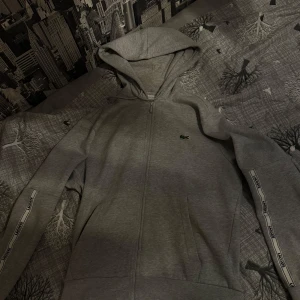 Grå hoodie från Lacoste med dragkedja - Snygg grå hoodie från Lacoste med dragkedja och huva. Klassisk passform, logga på bröstet och Lacoste-band längs ärmarna. Perfekt för chill dagar eller när du vill ha en clean streetstyle-look.