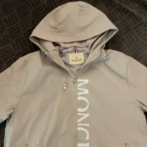 Grå vindjacka från Moncler med logga - Den är Storlek L men passar bara M och S och bäst för S skulle jag säga. Bra för hösten som kommer snart. Skulle säga att den passar bra med dem som inte har jätte lång överkropp och är runt 178 till 172 cm. Helt ny aldrig använd.  Snygg grå vindjacka från Moncler med stor vit logga längs dragkedjan och klassisk patch på ärmen. Jackan har huva, tre fickor med dragkedja och coola detaljer som Monclers signaturrand på insidan. Perfekt för dig som gillar stilren och sportig look. 
