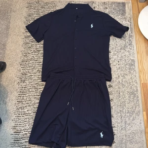 Mörkblå ribbad skjorta från Polo Ralph Lauren - Säljer en mörkblå ribbad skjorta från Polo Ralph Lauren med kort ärm och knappar framtill. Skjortan har klassisk krage och den ikoniska broderade loggan på bröstet. Materialet är mjukt och andas och polo Ralph lauren shortsen som matchar med skjortan, perfekt för varma dagar. Jag har aldrig använt dom förut för dom passade inte på mig. Pris kan diskuteras för än snabbare affär👍😃
