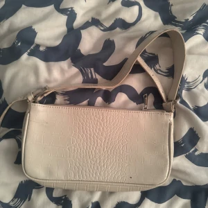Beige axelväska från Gina Tricot - Säljer en beige axelväska från Gina Tricot med croco-mönstrad yta och justerbart axelband. Väskan har dragkedja upptill och är i syntetmaterial. Perfekt storlek för det viktigaste och ger en clean vibe till din outfit.