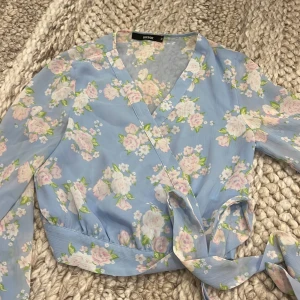Blommig från BikBok - Supersöt ljusblå blus från BikBok med blommigt mönster i vitt och rosa. Blusen har långa ärmar, v-ringning och knytband i midjan. Stängs med tryckknappar framtill. Storlek XS