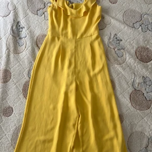 Gul byxdress från MNG i storlek XS - Snygg gul byxdress från MNG i storlek XS. Byxdressen har breda ben, ärmlös design och en volangdetalj vid halsringningen. Perfekt för dig som vill sticka ut med en färgglad och trendig look.