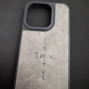 Cactus Jack iPhone 15 Pro skal - Snyggt iPhone 15 Pro skal med grå mockaliknande yta och svart ram. Framsidan har handskriven Cactus Jack-logga som ger en unik streetkänsla. Skalet har stor öppning för kameran och passar perfekt för dig som gillar Travis Scott-vibbar.