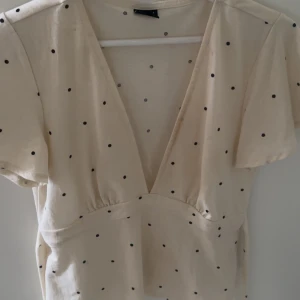 Prickig topp från Gina Tricot - Supersöt ljus topp från Gina Tricot med svarta prickar. Djup v-ringning och korta ärmar ger en trendig vibe. Perfekt för dig som gillar en feminin och lekfull stil.