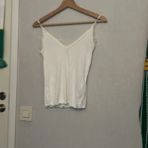 Vit spetslinnetopp från Amisu - Säljer en söt vit linnetopp från Amisu med tunna axelband och spetsdetaljer längs urringningen. Toppen har en lätt vågig kant nertill och är perfekt för lager-på-lager eller till jeans. Enkel och stilren design som funkar till många looks.