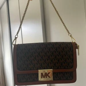 Michael Kors brun axelväska med kedja - Snygg brun axelväska från Michael Kors med guldiga detaljer och kedjerem. Väskan har MK-logga över hela ytan, magnetlås framtill och dragkedjefack inuti. Perfekt storlek för det viktigaste och ger en lyxig känsla till din outfit.
