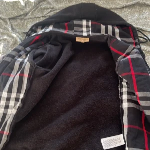 Svart fleecejacka från Burberry | S - Svart fleecejacka från Burberry med dragkedja framtill, hög krage med snörning och klassiskt rutigt foder i svart, vitt och rött. Jackan har broderad logga på bröstet och två fickor. Perfekt för dig som gillar stilren och bekväm design. Storlek S passar perfekt för 160-175