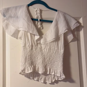 Vit blus med volang och knyt - Supersöt vit blus med bred volangkrage och smockad midja. Toppen har knyt i nacken, v-ringning och volangdetaljer längst ner. Perfekt för dig som gillar romantisk och luftig stil.