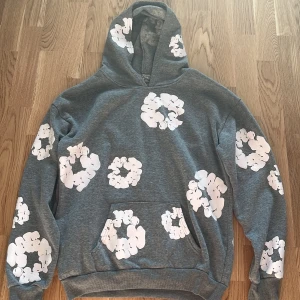 Grå hoodie från Denim Tears med blommor | S - Grå hoodie från Denim Tears med stora vita blommönster över hela tröjan. Klassisk känguruficka framtill och huva. Skön och avslappnad passform, perfekt för dig som gillar streetwear vibes. Storlek S sitter som M passar någon som är 170-180