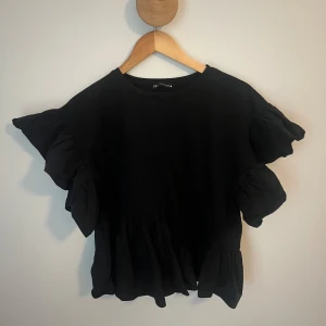 Svart volangblus med vida ärmar - Supersöt svart blus med volanger och vida korta ärmar. Toppen har en lös passform och volangdetalj nedtill. Perfekt att styla med jeans!! 