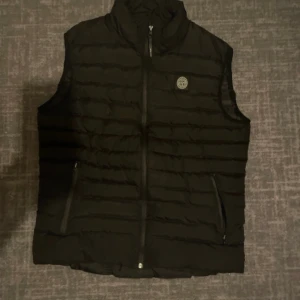 Svart dunväst från Stone Island - Säljer en svart dunväst från Stone Island med quiltad design och hög krage. Västen har dragkedja framtill, två fickor med dragkedja och en snygg logotyp på bröstet. Perfekt att slänga över en hoodie för en clean och sportig look. Fickan är dock lite sönder på insidan