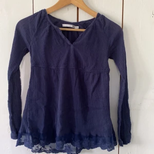 Mörkblå blus med spets från 157 - Supersöt mörkblå blus från 157 med v-ringning och långa ärmar. Blusen har en flowy passform och är dekorerad med fin spetsdetalj längst ner. Perfekt för dig som gillar en romantisk och chill stil.