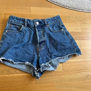 Blå högmidjade jeansshorts H&M - Snygga blå jeansshorts från H&M med hög midja och råa, fransiga kanter. Klassisk femficksmodell i denim med vintagekänsla och slitna detaljer. Perfekta för sommaren och ger en avslappnad vibe till din outfit.