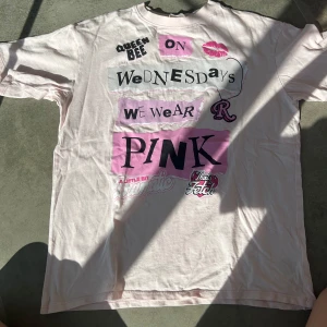 Rosa t-shirt med tryck från H&M - Ljusrosa t-shirt från H&M med coolt tryck inspirerat av Mean Girls. Texten 'On Wednesdays we wear pink' och flera grafiska detaljer i svart och rosa. Klassisk passform med korta ärmar och rund hals. Perfekt statement-plagg för dig som gillar popkultur.