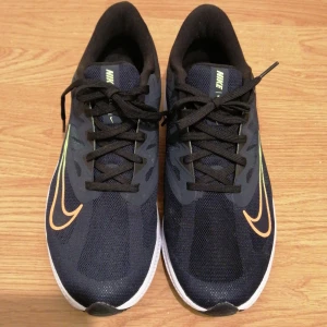 Nike löparskor marinblå strl 44 - Snygga marinblå Nike löparskor med svart snörning och vit sula. Skorna har mesh-ovandel för bra andning och en stor Swoosh-logga i orange och limegrönt på sidan. Perfekta för dig som gillar att springa och vill ha stilrena sneakers med sportig vibe.