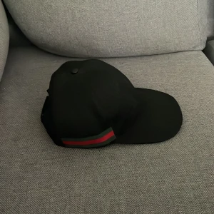 Svart keps med röd och grön rand - Snygg svart keps från Gucci med klassisk böjd skärm och justerbar rem baktill. Kepsen har en ikonisk röd och grön rand på sidan som ger en exklusiv känsla. Perfekt accessoar för att lyfta din streetwear-look.