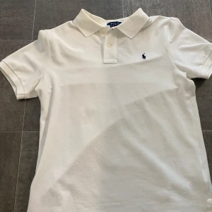Vit pikétröja från Polo Ralph Lauren - Klassisk vit pikétröja från Polo Ralph Lauren i slim fit-modell. Tröjan har korta ärmar, krage med knappar och den ikoniska broderade loggan i mörkblått på bröstet. Tillverkad i bomull för en skön och luftig känsla.