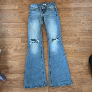 Bootcut jeans från Nelly med slitningar - Ljusblå bootcut jeans från Nelly med slitna detaljer på knäna och är lågmidjade! Klassisk femficksmodell med dubbla knappar och dragkedja. Jeansen har en snygg utsvängd passform. Byxorna har dock blivit nötta längst på högra benat på grund utav att byxorna har varit för långa på mig!