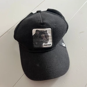 Svart keps med panterpatch Goorin Bros - Svart keps från Goorin Bros med en broderad patch framtill som visar en panter och texten 'Panther'. Kepsen har klassisk böjd skärm och mesh baktill för extra ventilation. Snygg detalj med varumärkets logga på sidan. Nypris 450kr. 