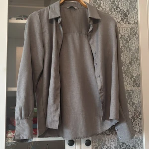 Grå långärmad skjorta i linne - Stilren grå skjorta i linne med klassisk krage och knappar framtill. Skjortan har långa ärmar med dubbla knappar vid manschetten och en rak passform. Perfekt för en clean och avslappnad look.