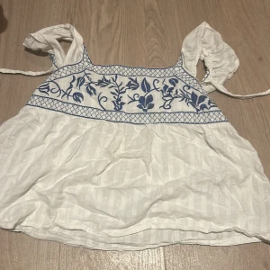 Vit blommig linnetopp med blå broderi - Supersöt vit linnetopp med blått broderat blommönster upptill och knytband i ryggen. Toppen har breda axelband och lätt volangig nederdel. Perfekt för varma dagar och ger en fräsch sommarkänsla.