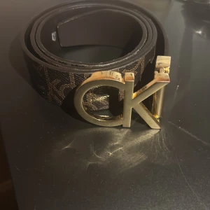 Brunt CK bälte med guldspänne - Snyggt brunt bälte från Calvin Klein med stort guldigt CK-spänne. Bältet har ett diskret mönster med CK-loggor längs hela remmen och är gjort i skinn. Perfekt accessoar för att lyfta din outfit med en lyxig touch.