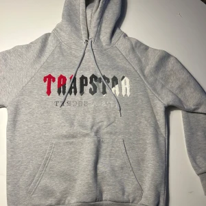 Grå hoodie Trapstar med broderad text - Säljer en grå hoodie från Trapstar med broderad text i rött, svart och vitt på bröstet. Klassisk känguruficka, huva med snörning och mjukt material. Perfekt för dig som gillar streetwear och vill ha en clean look.