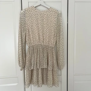 Prickig beige kortklänning med volang - Supersöt beige kortklänningfrån Gina tricot i storlek 36 med svarta prickar och långa ärmar. Klänningen har volangig kjol 