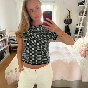 Svartvit mönstrad topp med knapp i nacken - Säljer en svart topp med vita prickar och korta ärmar. Toppen har en rund halsringning med svart kant och en snygg knappdetalj i nacken. Modellen är figurnära och croppad, perfekt till höga jeans eller kjol.