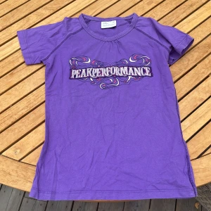Lila Peak Performance t-shirt junior - Snygg lila t-shirt från Peak Performance i juniorstorlek 140. T-shirten har korta ärmar och ett coolt tryck med färgglada detaljer och logga framtill.  Skön och mjuk bomullskvalitet.