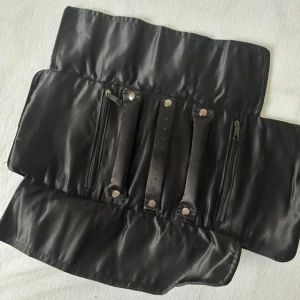 Svart smyckesväska från Dyrberg/Kern - Snygg svart smyckesväska från Dyrberg/Kern. Väskan rullas ihop och knyts med ett band med logga. Inuti finns flera fack med dragkedja och remmar med tryckknappar för att hålla smycken på plats. Perfekt för att organisera dina favoriter på resan