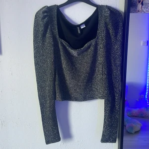 Glittrig långärmad topp Divided S - Snygg glittrig svart topp från Divided med croppad passform och långa ärmar. Toppen har lätt puffade axlar och en rundad urringning. Perfekt för dig som vill sticka ut med lite extra glitter. Materialet är stretchigt och bekvämt.